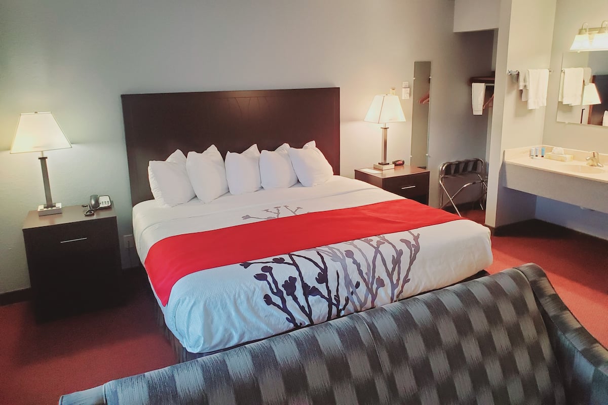 A hotel room with a large bed, two bedside tables with lamps, a chair, a sofa, and a bathroom with a sink and mirror.