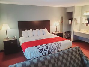 A hotel room with a large bed, two bedside tables with lamps, a chair, a sofa, and a bathroom with a sink and mirror.