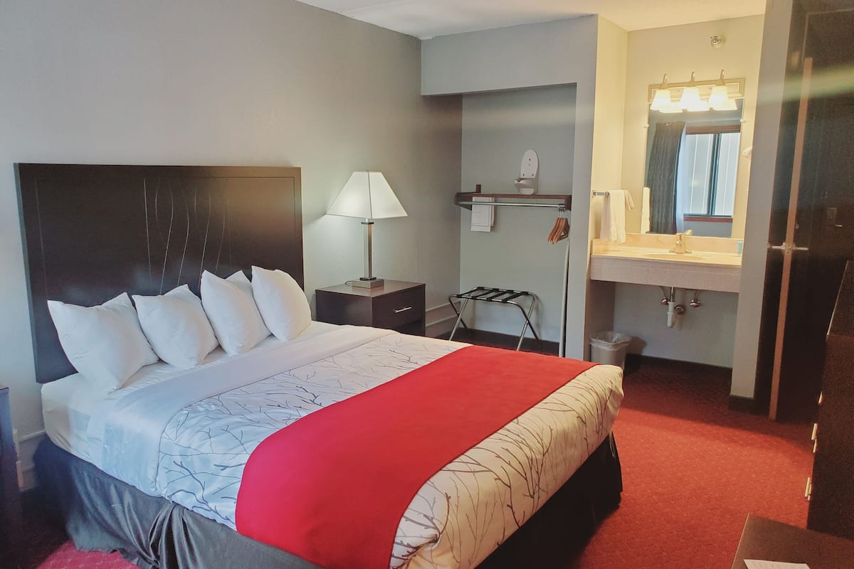 A hotel room with a large bed, a nightstand with a lamp, a small table, a mirror, and a bathroom with a sink and towel rack.