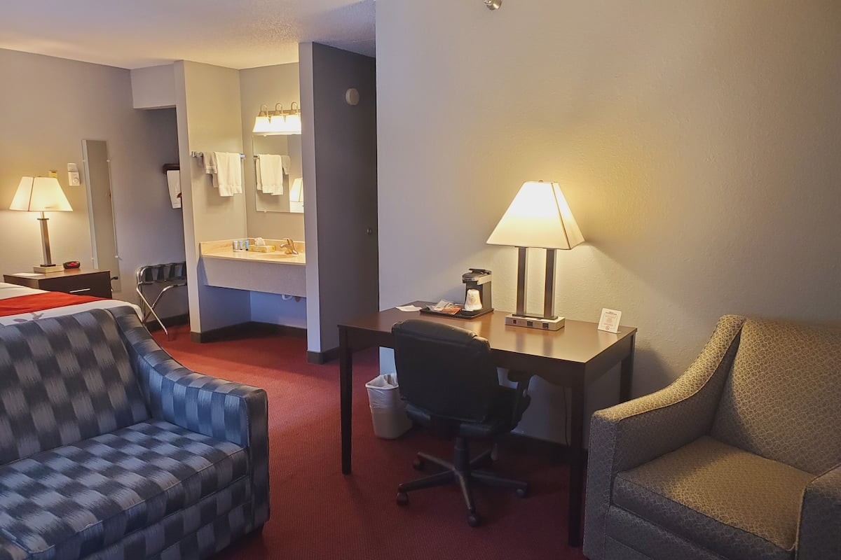 A hotel room with a checkered sofa, a desk with a lamp, a chair, a small table, a trash bin, and a bathroom with a mirror and towel rack.