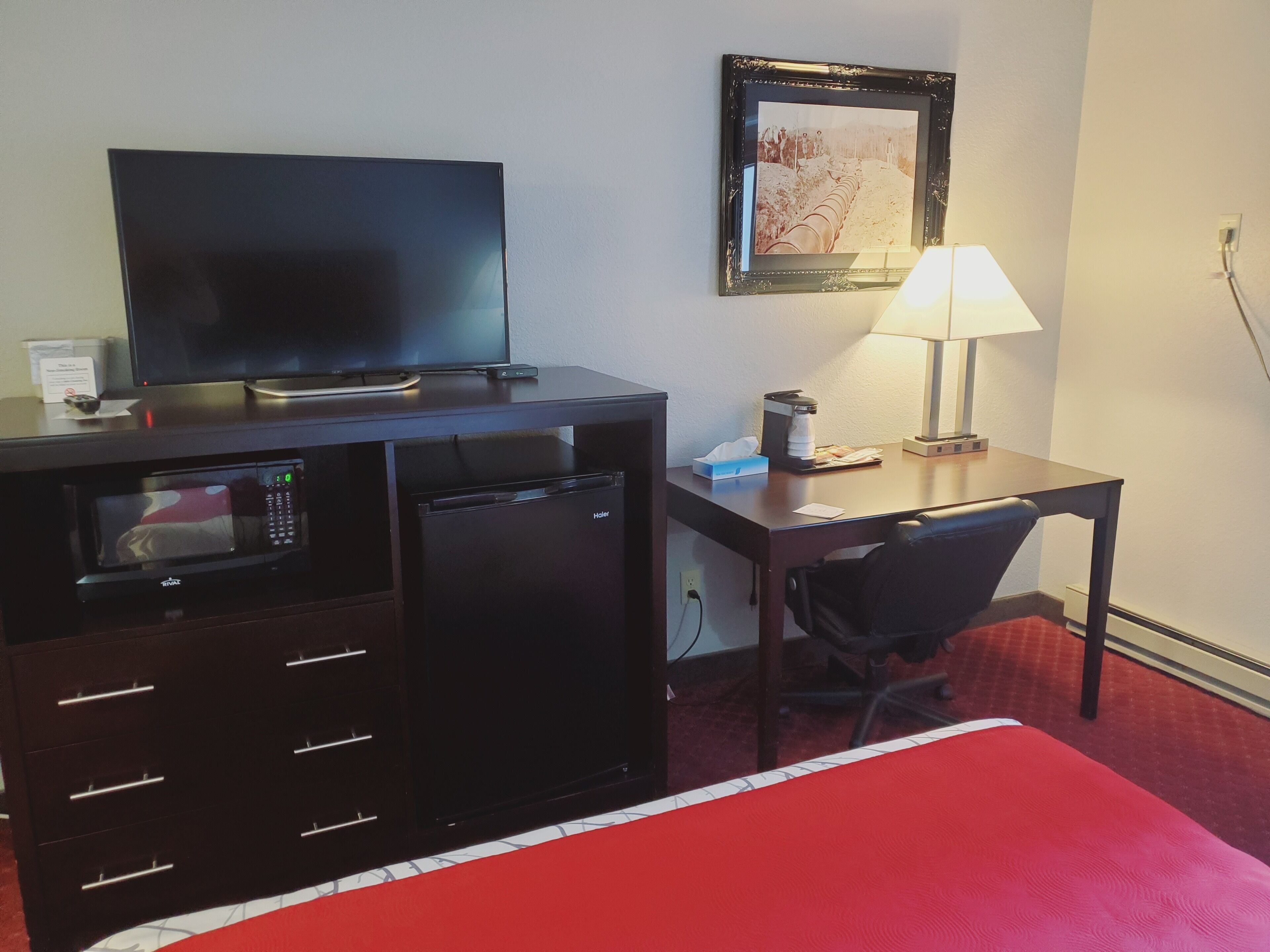 Een hotelkamer met een bed, een bureau met lamp, een televisie, een magnetron en een koffiezetapparaat.