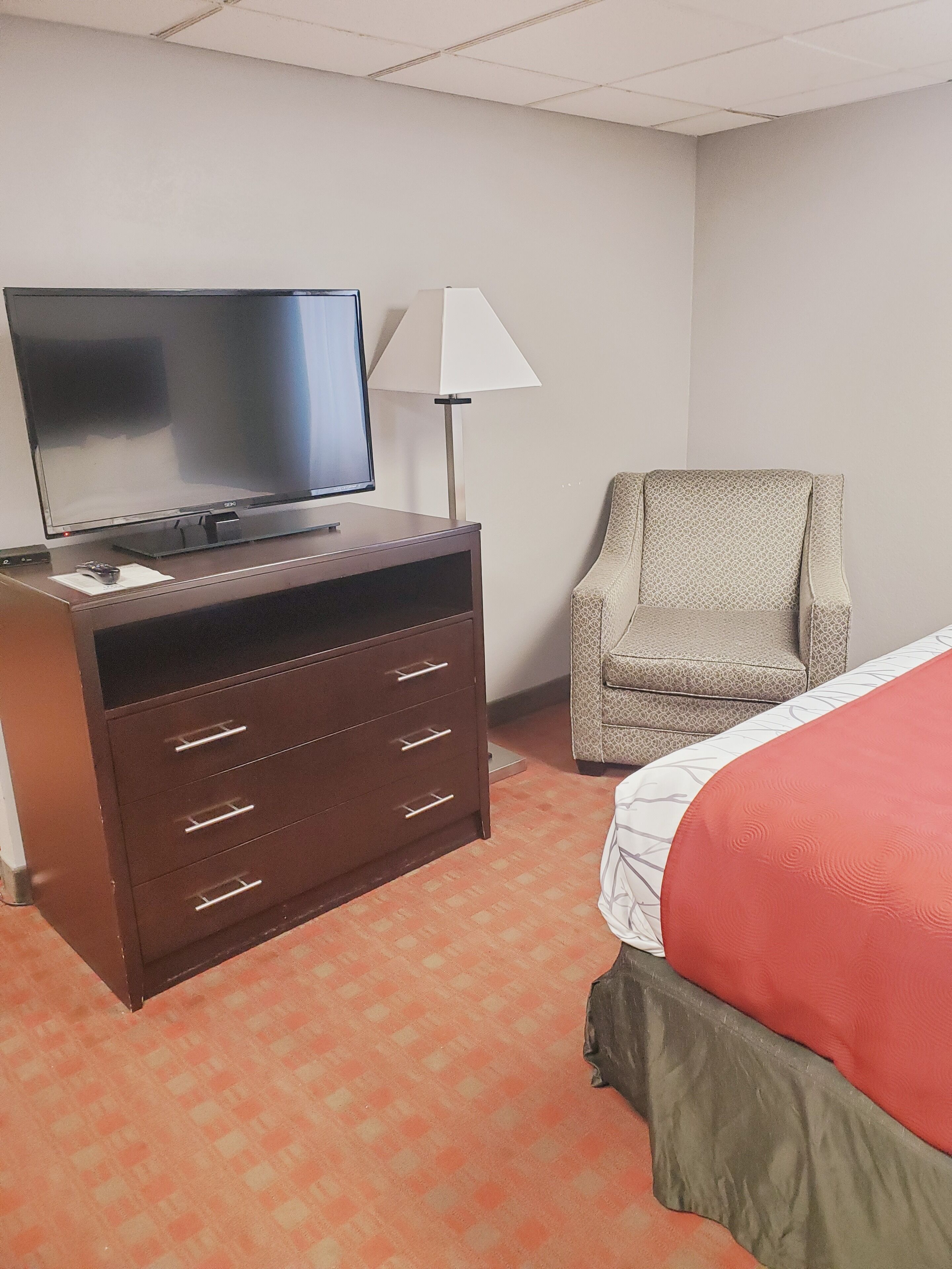A hotel room with a flat-screen TV on a wooden dresser, a patterned armchair, and a bed with a red bedspread.