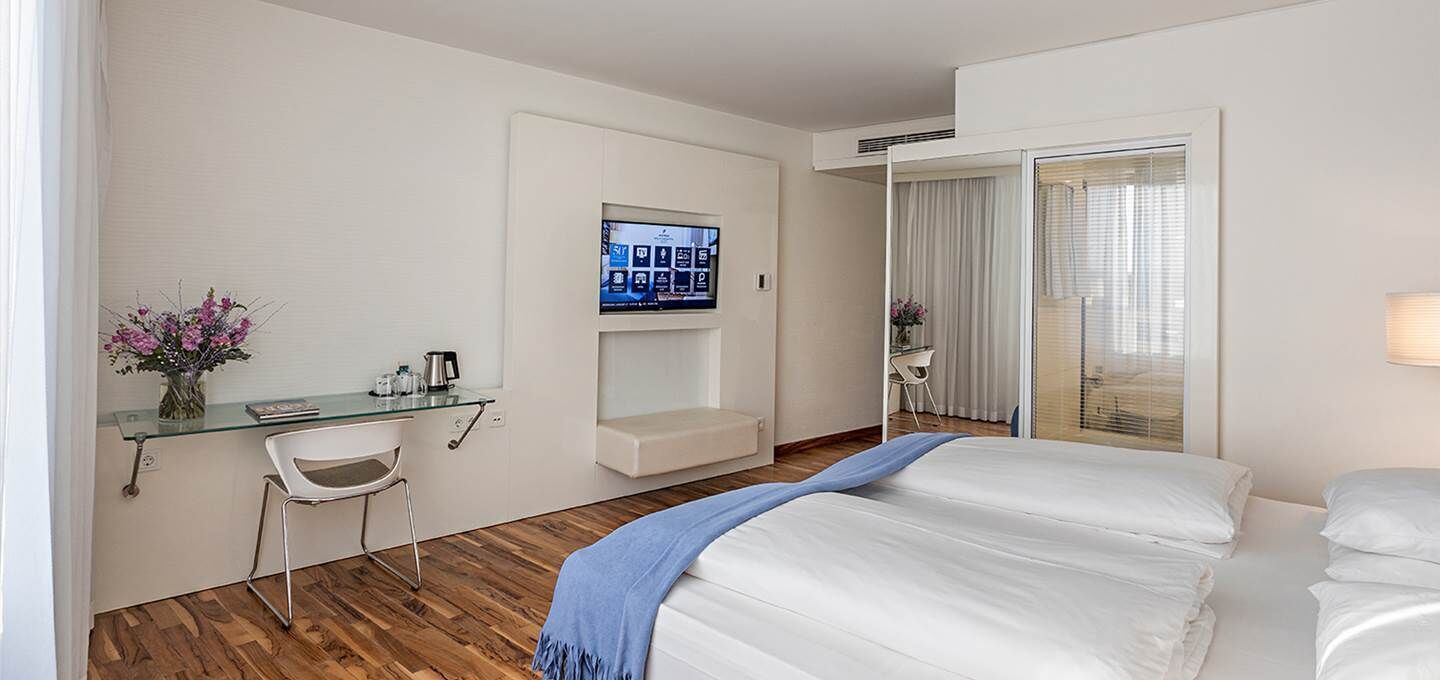Habitación de hotel con cama, escritorio con monitor de computadora, una silla y una mesita con un jarrón de flores.