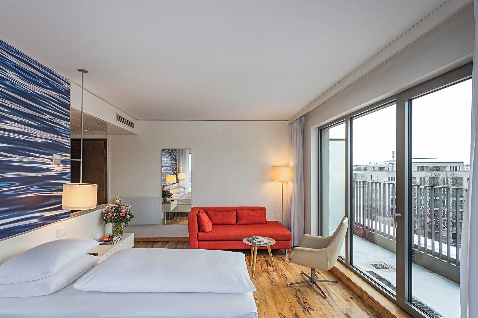 Ein Hotelzimmer mit großem Fenster, einem Bett, einem roten Sofa, einem Sessel und einem Spiegel.