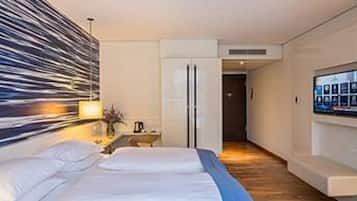 A modern hotel room with a large bed, a flat-screen TV mounted on the wall, and a wooden floor.