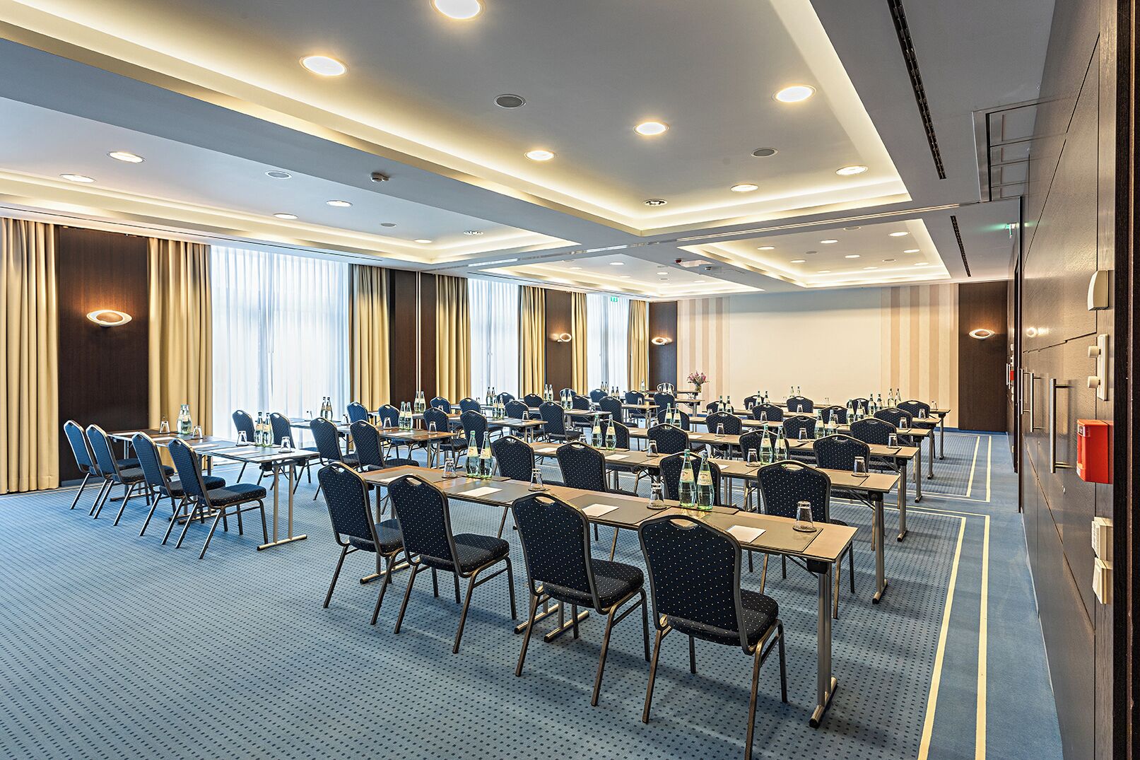 Ein Konferenzraum mit mehreren Tischen und Stühlen, großen Fenstern mit Vorhängen und einem modernen Deckenstil.