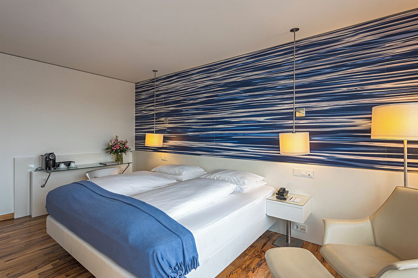 Una camera d'albergo moderna con un letto grande, lampade sui comodini, un tavolino con il telefono e una sedia.