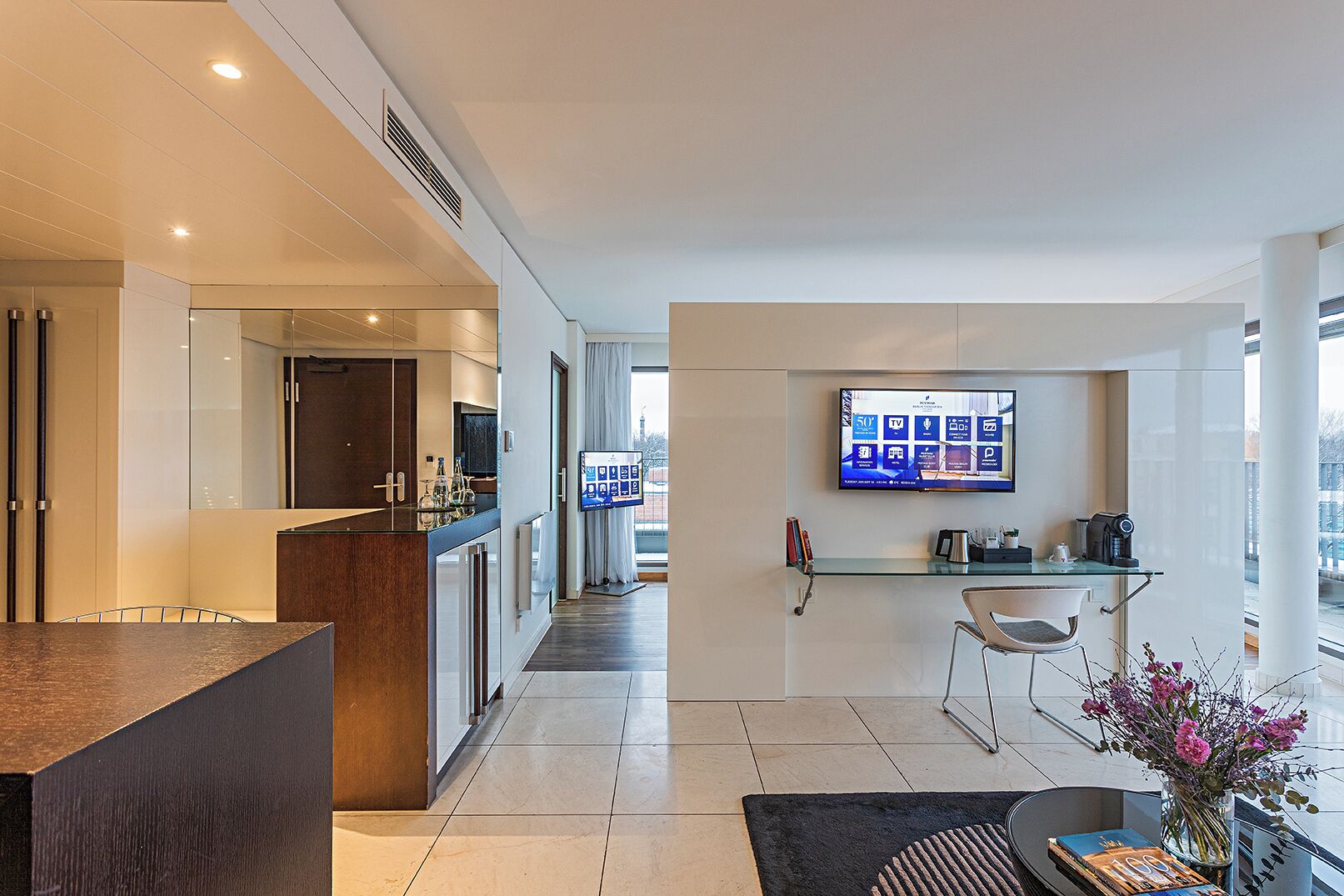 A modern hotel room with a kitchen area, a dining space, and a large TV mounted on the wall.