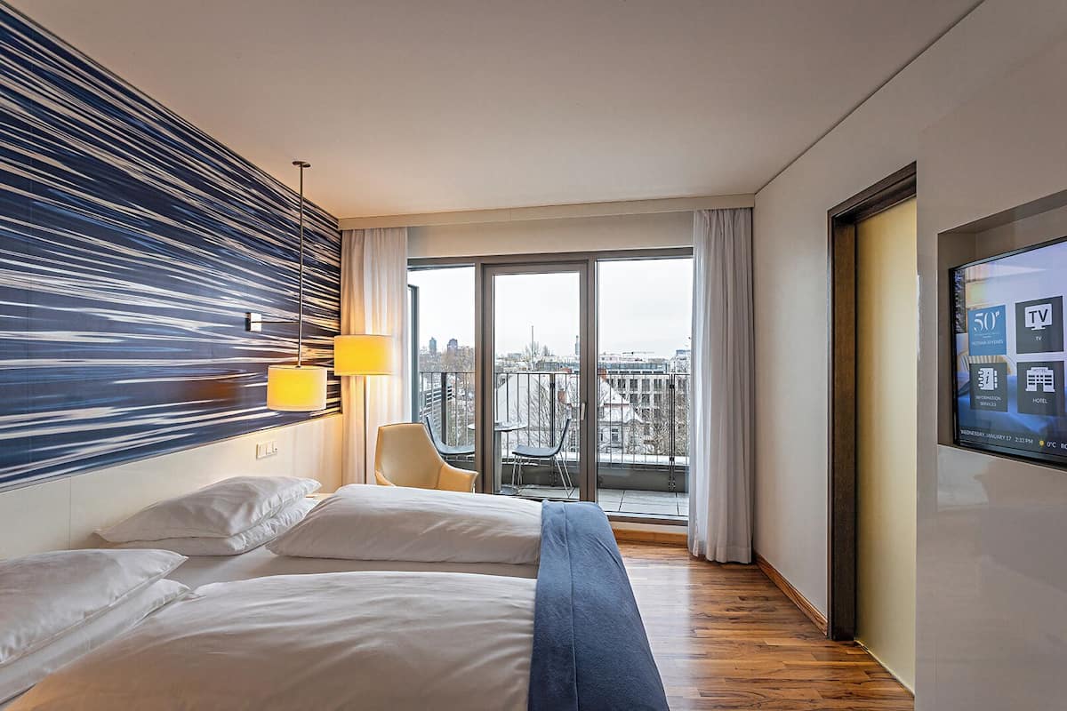 A hotel room with two beds, a large window with a city view, a TV on the wall, and a modern lamp.