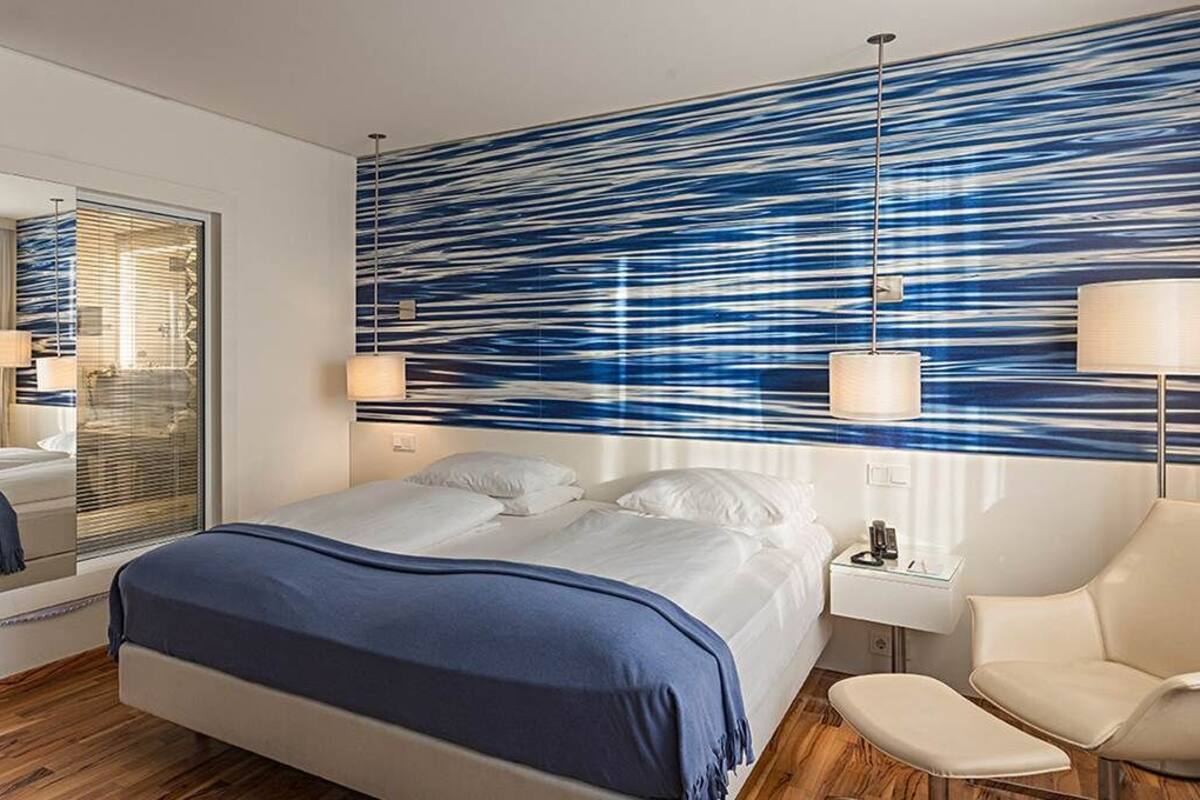 A modern hotel room with a large bed, a seating area with a chair and a small table, a large mirror, and a wall with a blue abstract design.
