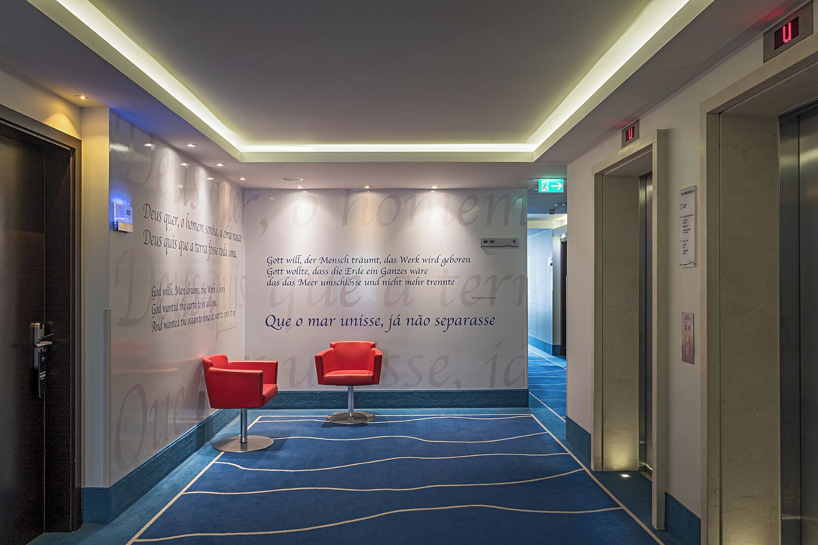 Ein Hotelgang mit blauem Teppich, roten Stühlen und einer Wand mit deutschem Text.