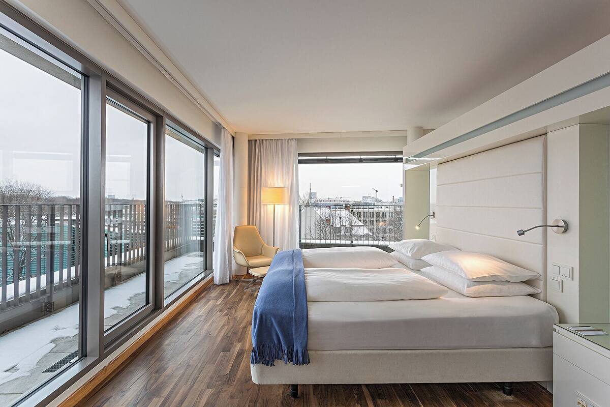 A modern hotel room with a large window, a bed, a chair, and a bedside table.