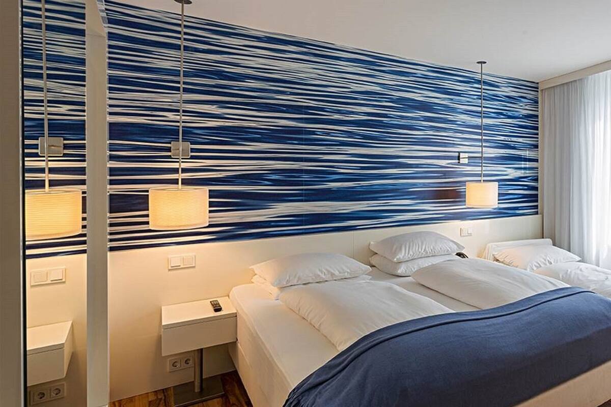 A hotel room with two beds, a modern blue and white wall design, a large window with sheer curtains, and a wooden floor.