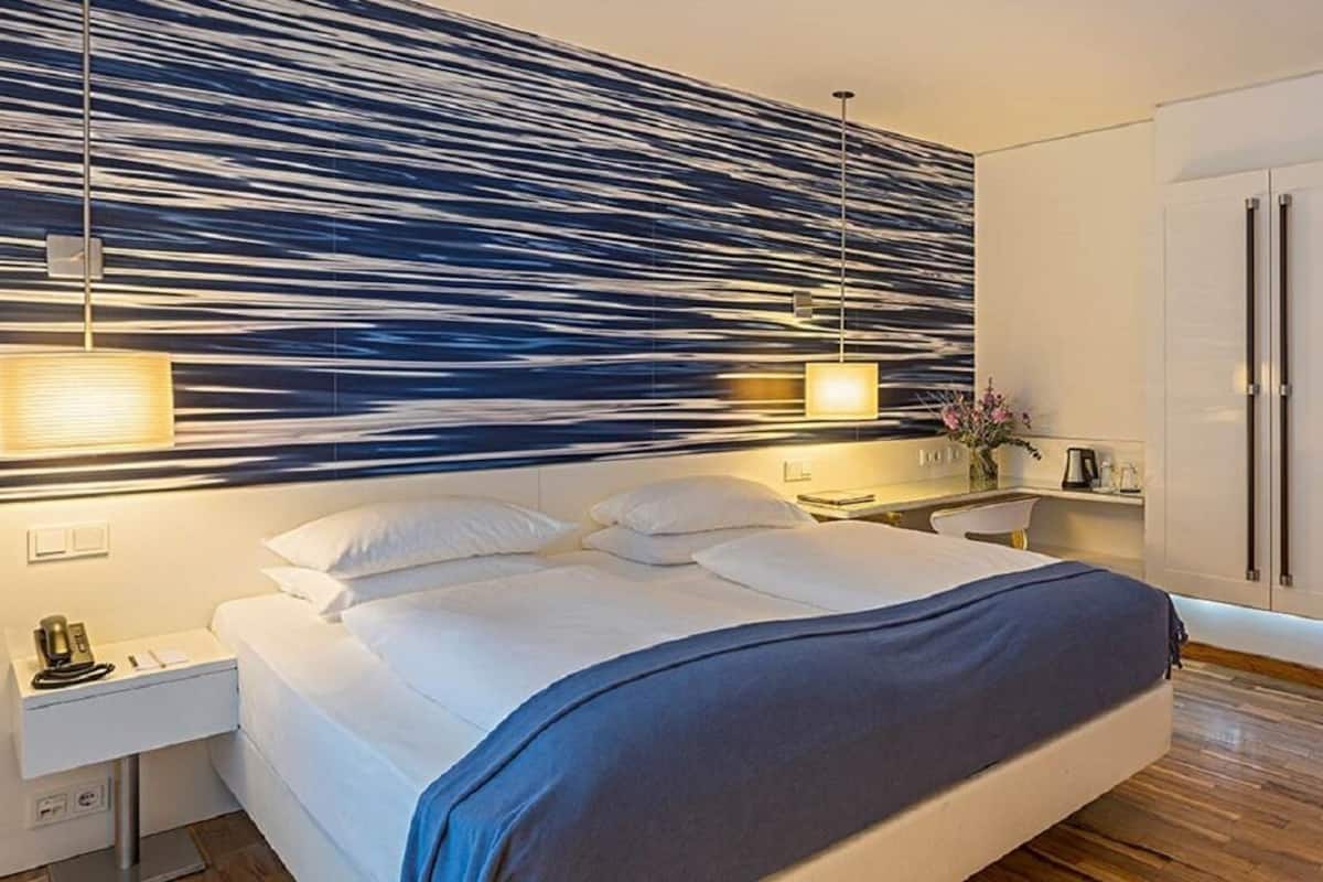 A modern hotel room with a large bed, bedside tables, a wardrobe, and a wall with a unique light pattern.