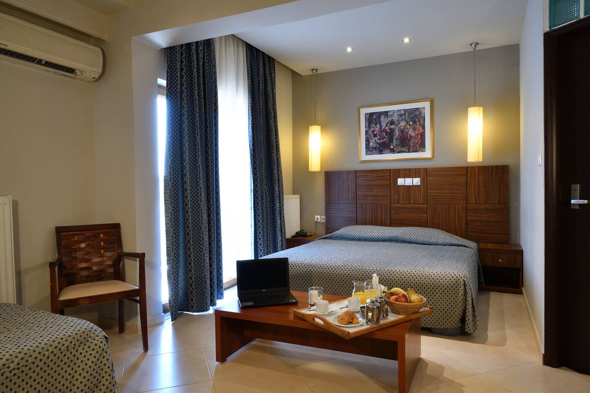 A hotel room with a bed, a desk with breakfast, a chair, and a painting on the wall.