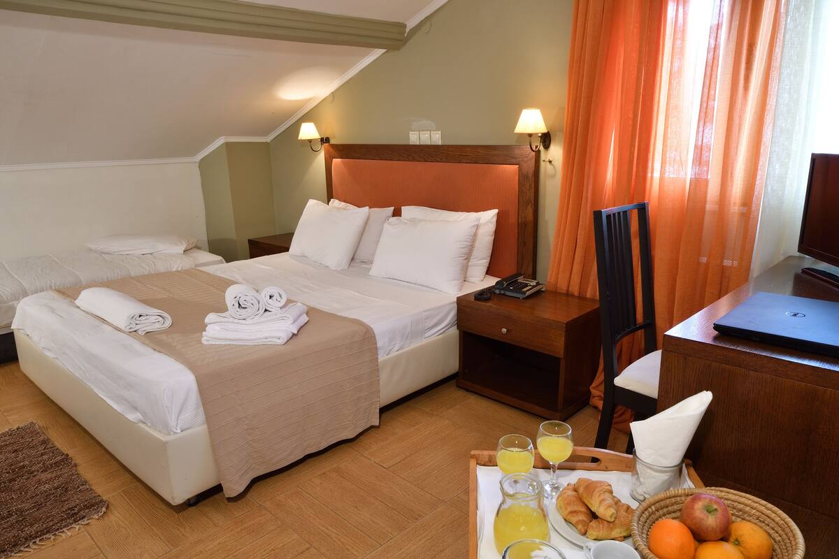 A hotel room with two beds, a desk with a laptop, a chair, and a breakfast tray with croissants, juice, and fruit.