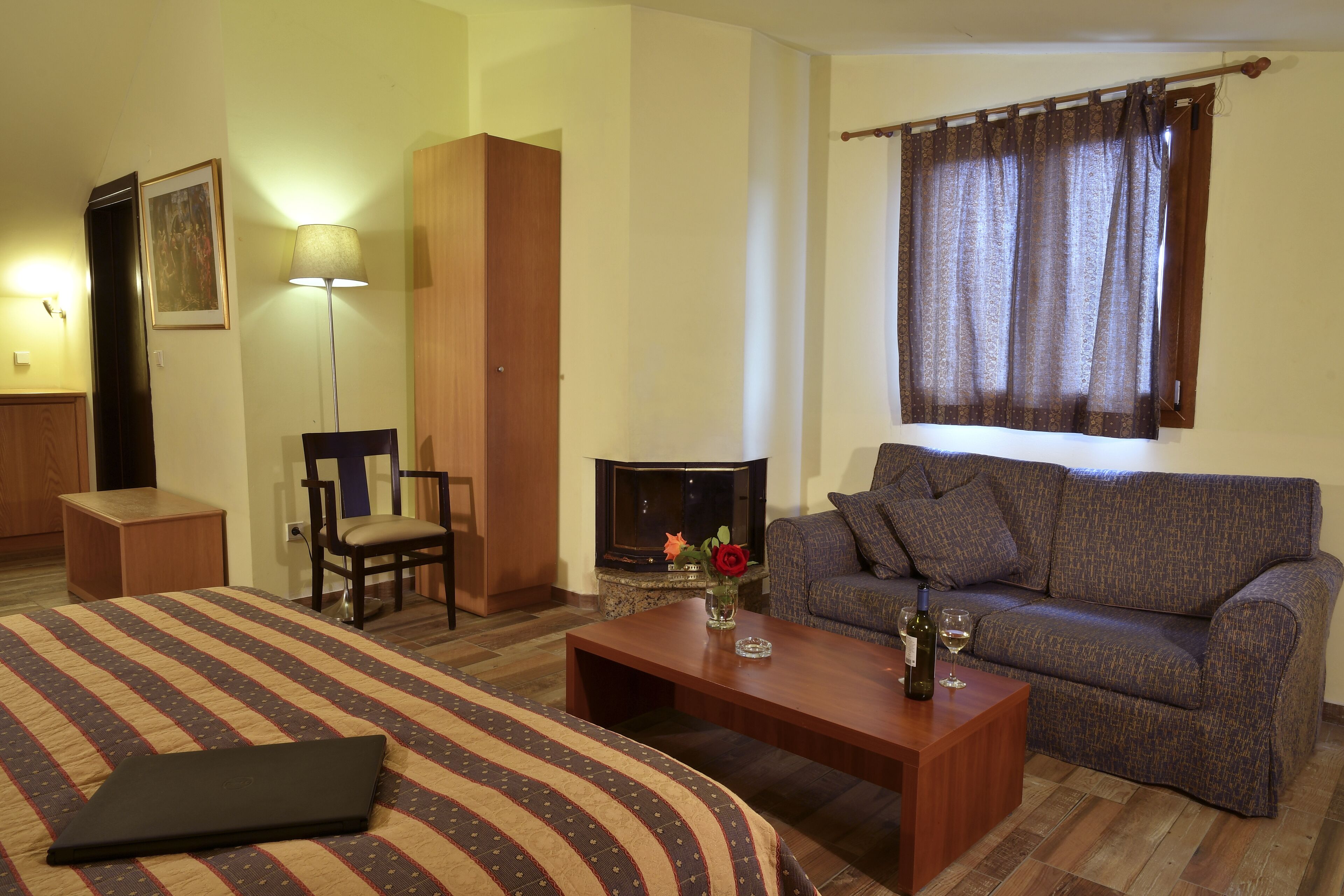Quarto de hotel com cama, sofá, mesa de centro, luminária, poltrona, lareira e janela com cortinas.