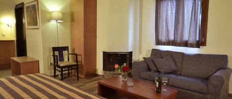 Quarto de hotel com cama, sofá, mesa de centro, luminária, poltrona, lareira e janela com cortinas.