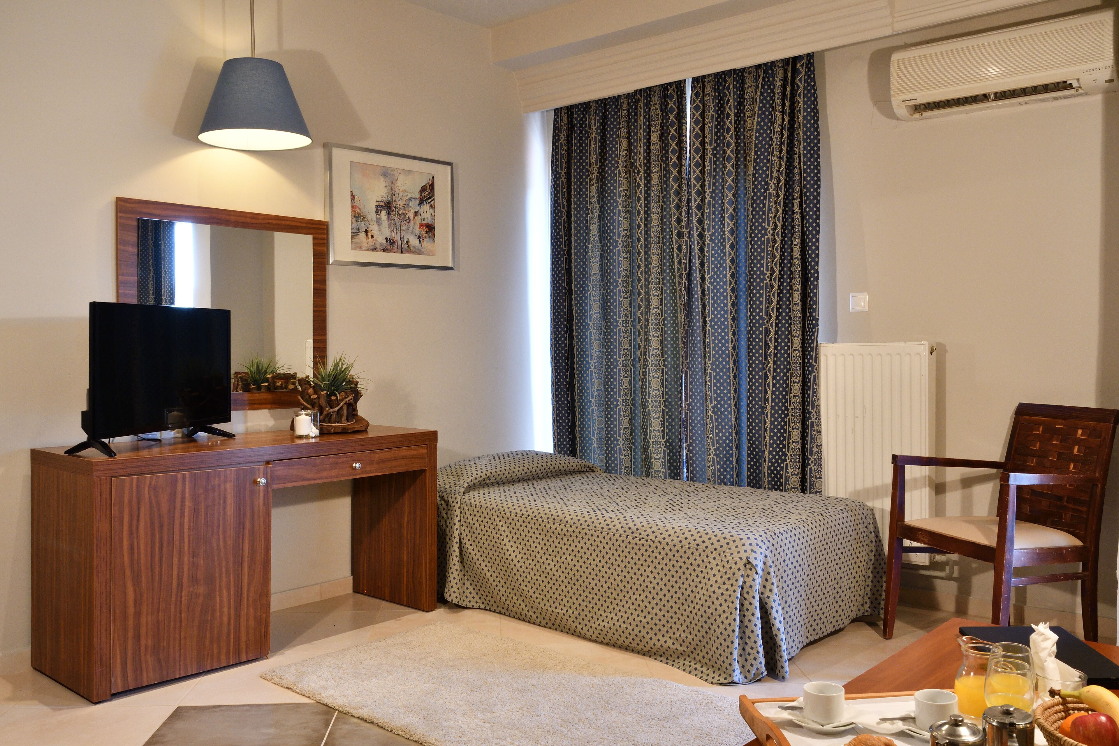 Ein Schlafzimmer mit Bett, Schreibtisch mit Fernseher, Stuhl, Fenster mit Vorhängen und Heizkörper.
