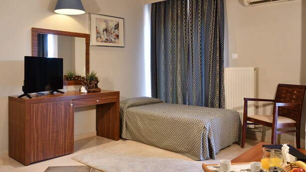 A bedroom with a bed, a desk with a TV, a chair, a window with curtains, and a radiator. - Four Seasons Hotel (Thermi)
