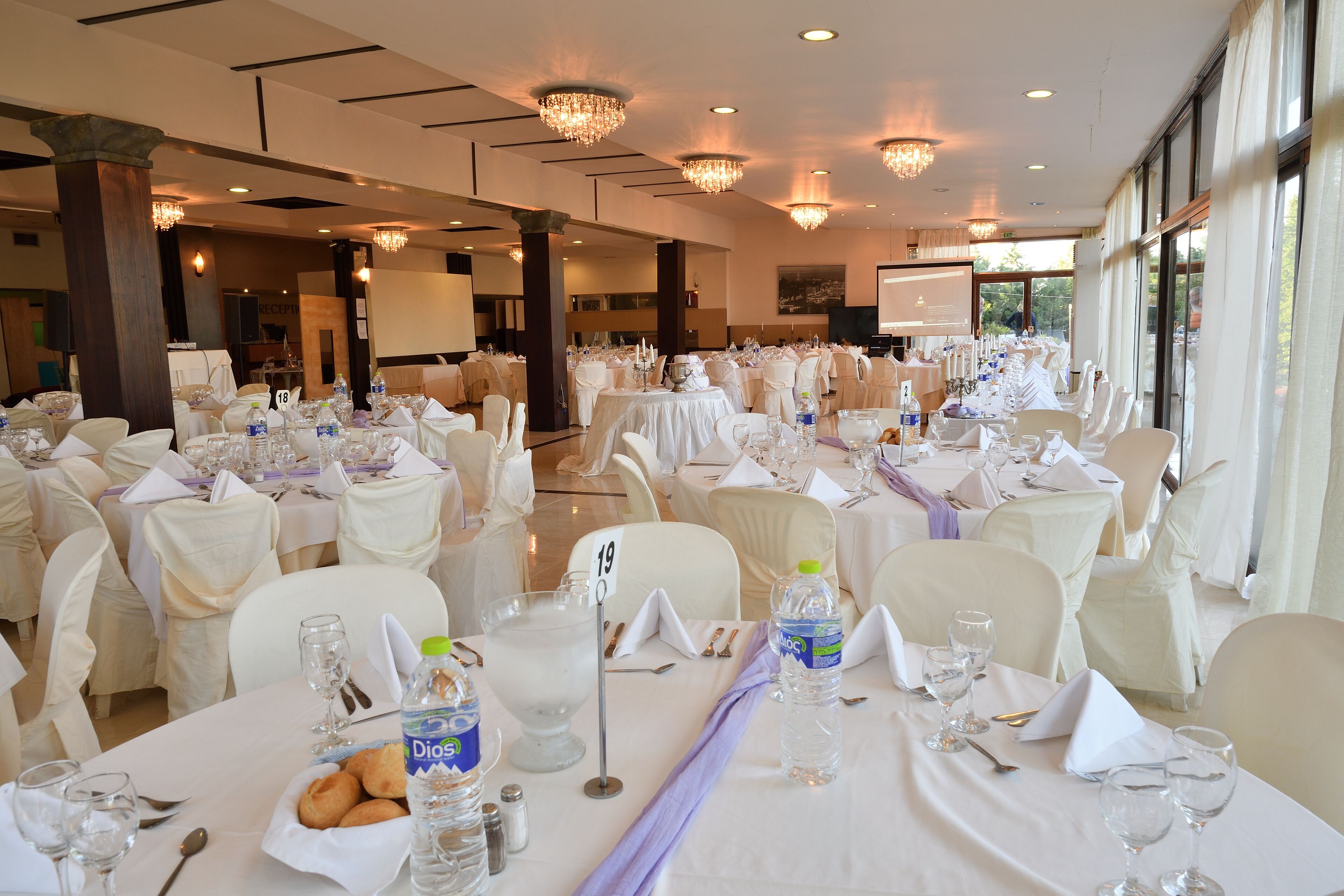 Een balzaal met ronde tafels gedekt voor een formeel evenement, met witte tafellakens, stoelen en middendeuren.