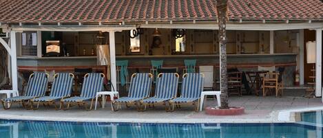 A pool area with lounge chairs, a covered seating area, and a building with a tiled roof.
