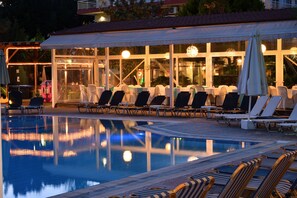 Un área junto a la piscina con sillones de descanso y sombrillas, una zona de estar cubierta y un reflejo en el agua.