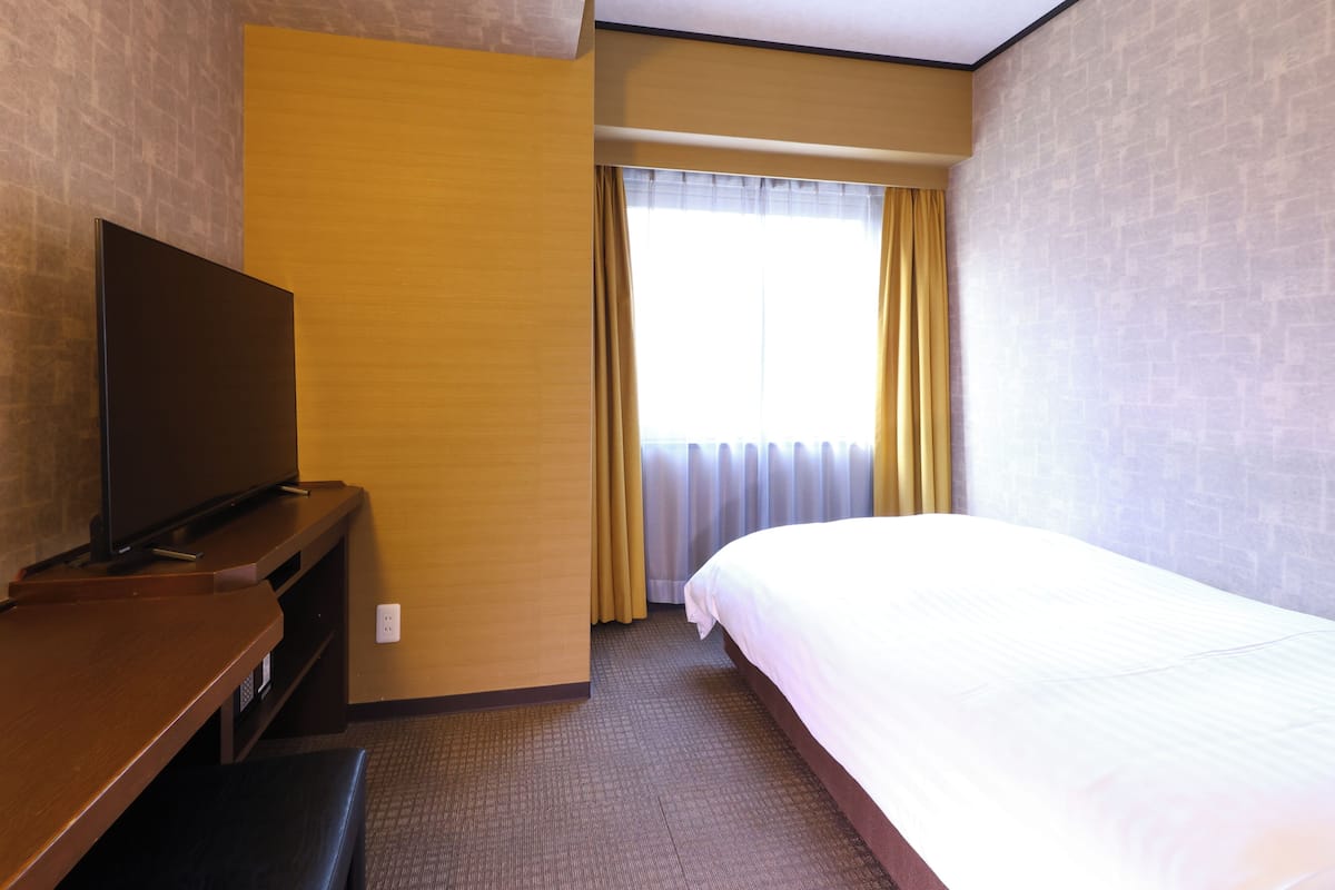 A hotel room with a bed, a television on a wooden stand, a window with curtains, and a carpeted floor.