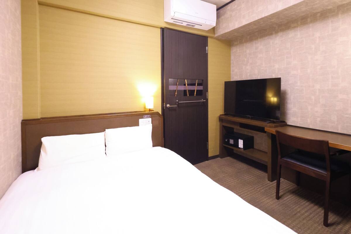 A hotel room with a bed, a desk, a television, and an air conditioning unit.