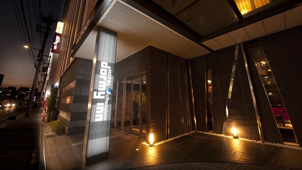 En modern hotellentrén med en glasfasad, en synlig receptionsdisk och en väl upplyst gångväg som leder fram till entrén.
