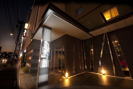 A modern hotel entrance with a glass facade, a visible reception desk, and a well-lit pathway leading to the entrance.