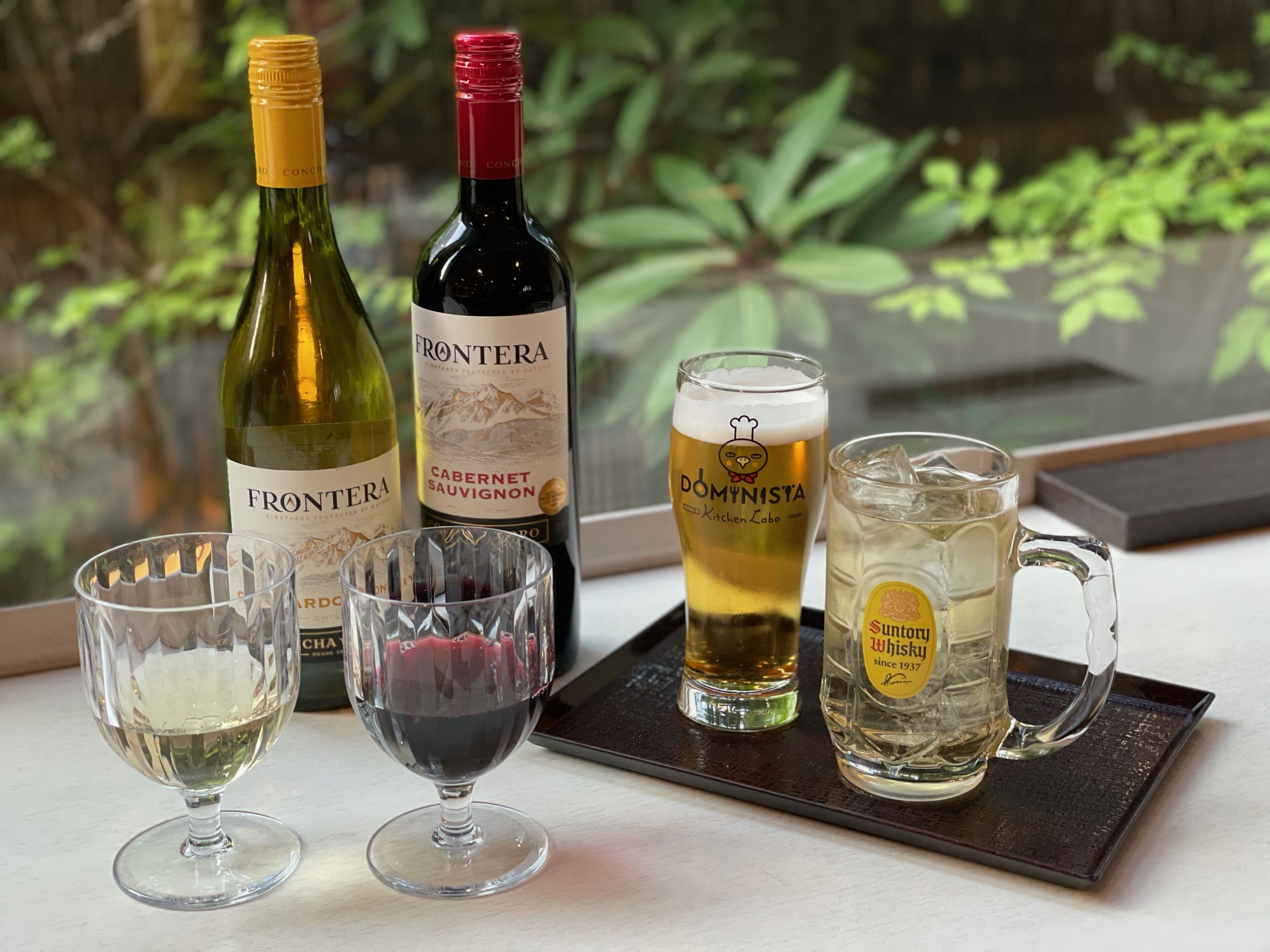 Une sélection de verres à vin et de verres à bière, une bouteille de vin et une autre bouteille de vin, le tout disposé sur un plateau près d’une fenêtre donnant sur un paysage verdoyant.