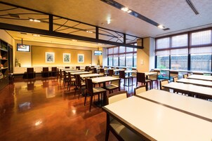 A spacious dining area with multiple tables and chairs, large windows with blinds, and artwork on the walls.