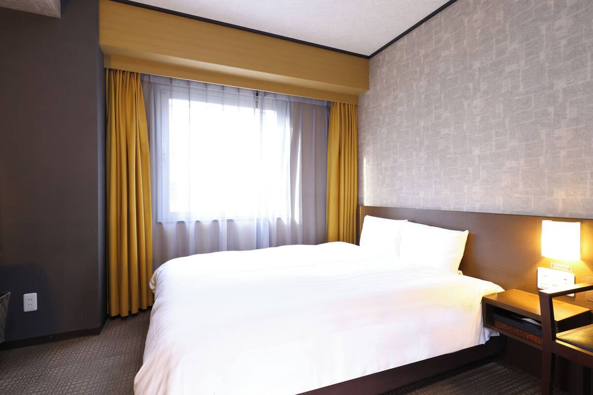 A hotel room with a bed, a nightstand, a lamp, a window with curtains, and a wall with a textured pattern.