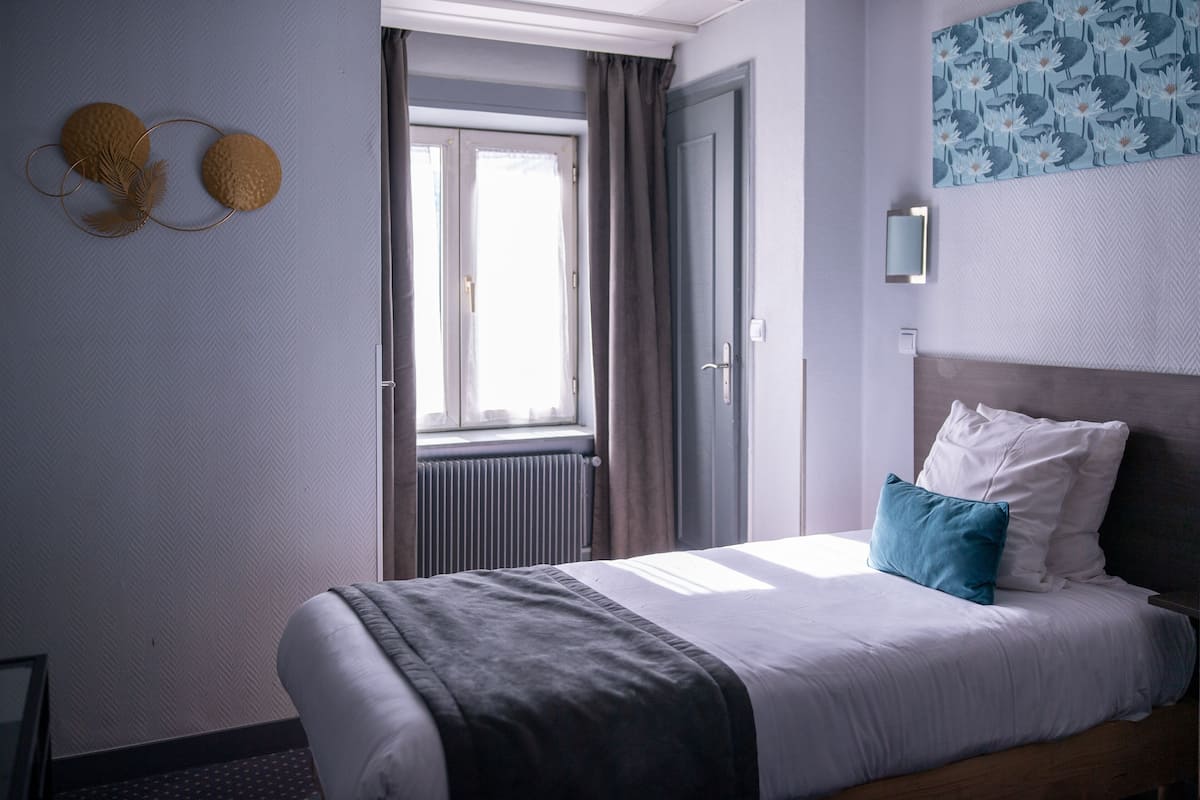 Una habitación de hotel con cama, ventana, radiador y lámpara fijada a la pared.