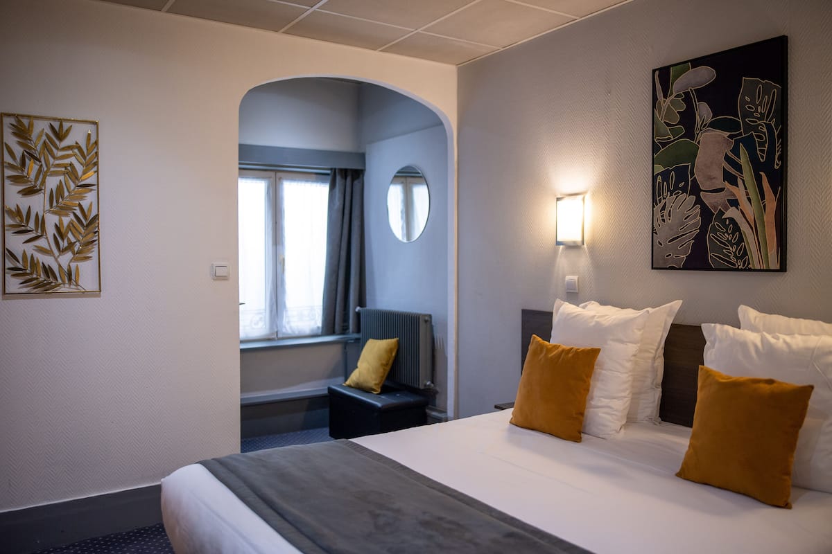 Habitación de hotel con cama, zona de estar con una silla y una mesa, un ventanal grande y obras de arte en las paredes.