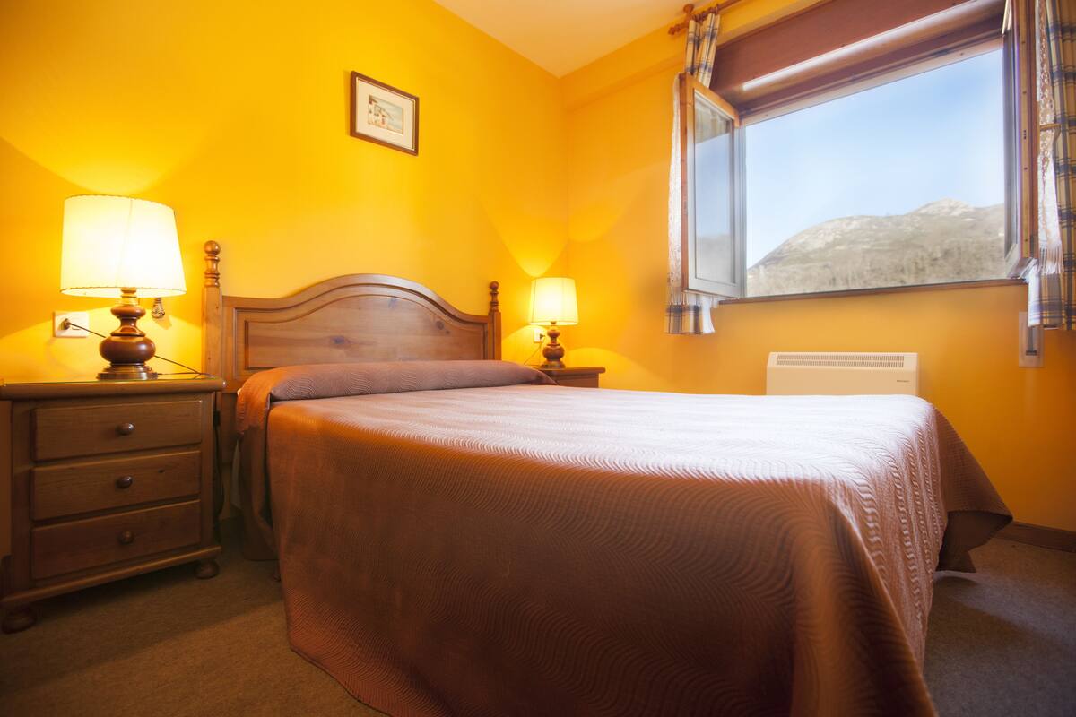 Un dormitorio con una cama, dos mesitas de noche con lámparas, un radiador y una ventana con cortinas.