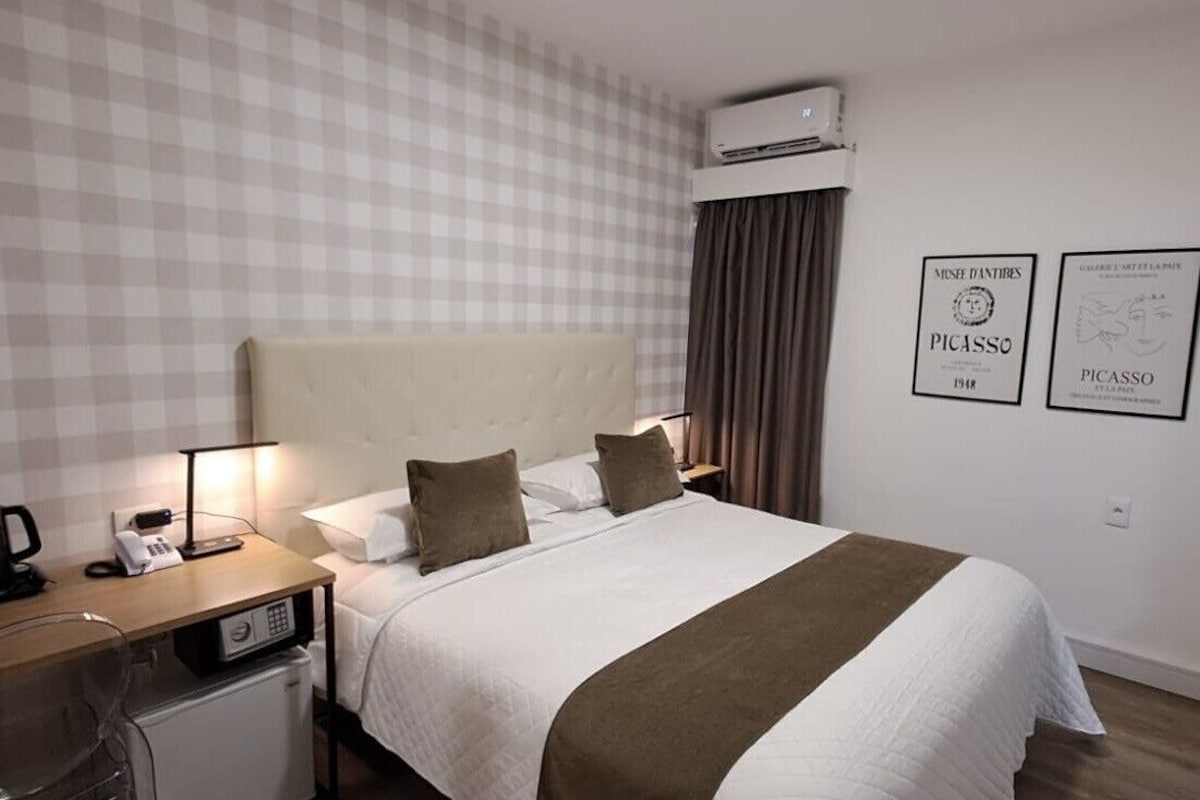 A hotel room with a checkered wall, a bed with a white and brown bedspread, a bedside table with a lamp, a small fridge, and two framed artworks on the wall.