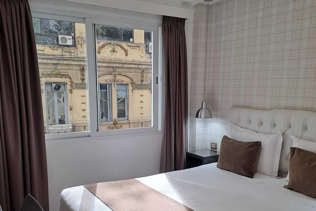 A hotel room with a large bed, a view of a building through the window, and a headboard with pillows.