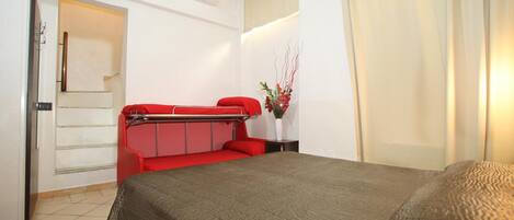 Una habitación de hotel con una cama, un sofá rojo, un televisor montado en la pared y una ventana con cortinas.