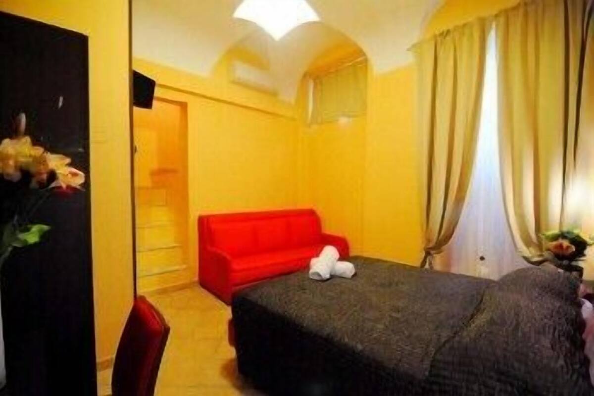 A room with a bed, a red sofa, and a small table with a vase of flowers.