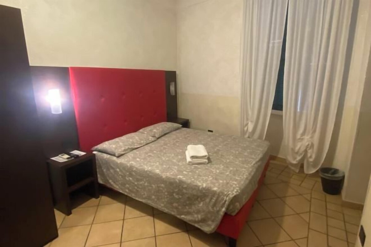 A bedroom with a bed, a nightstand, a red headboard, a white curtain, and a tiled floor.