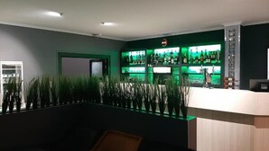 A modern bar area with a green-lit shelf displaying various bottles, a reception desk, and a seating area with a sofa and plants. - Hôtel Martinez Grenay (Grenay)