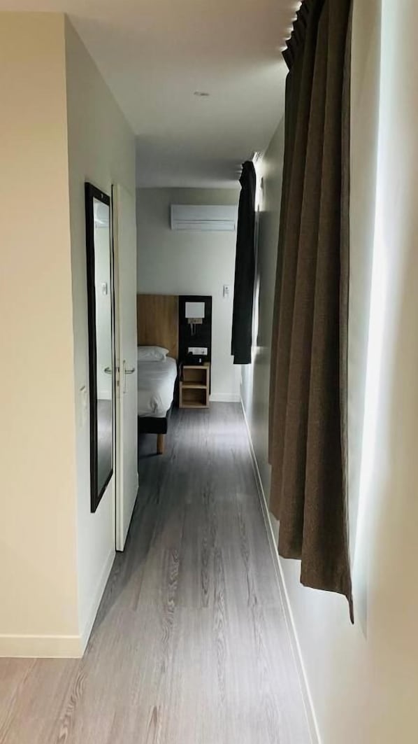 A narrow hallway with a bed at the end, a wooden floor, and a window with curtains.