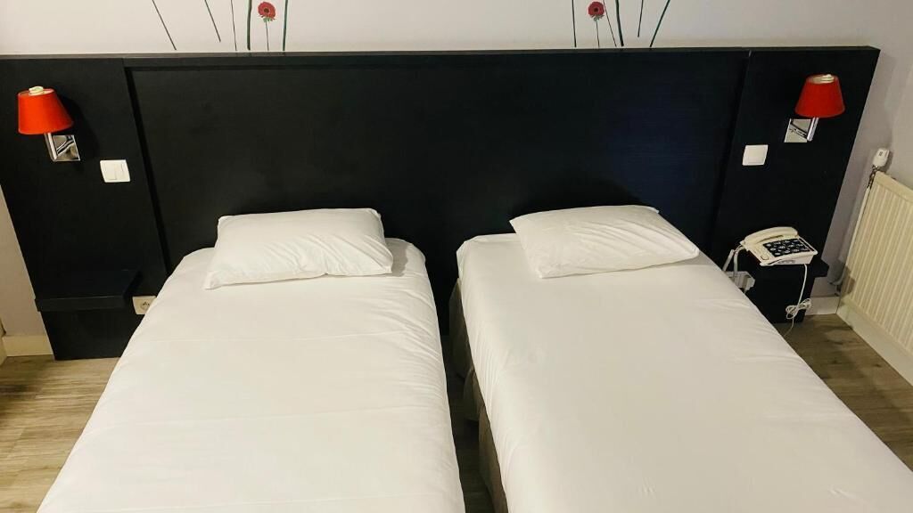 Ein Hotelzimmer mit zwei Einzelbetten, einem schwarzen Kopfteil, roten Wandlampen, einem weißen Telefon und einem Heizkörper.