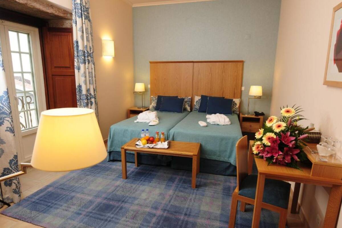 A hotel room with two beds, a wooden table with a tray of fruits and bottles, a chair, a desk with a vase of flowers, and a large lamp.
