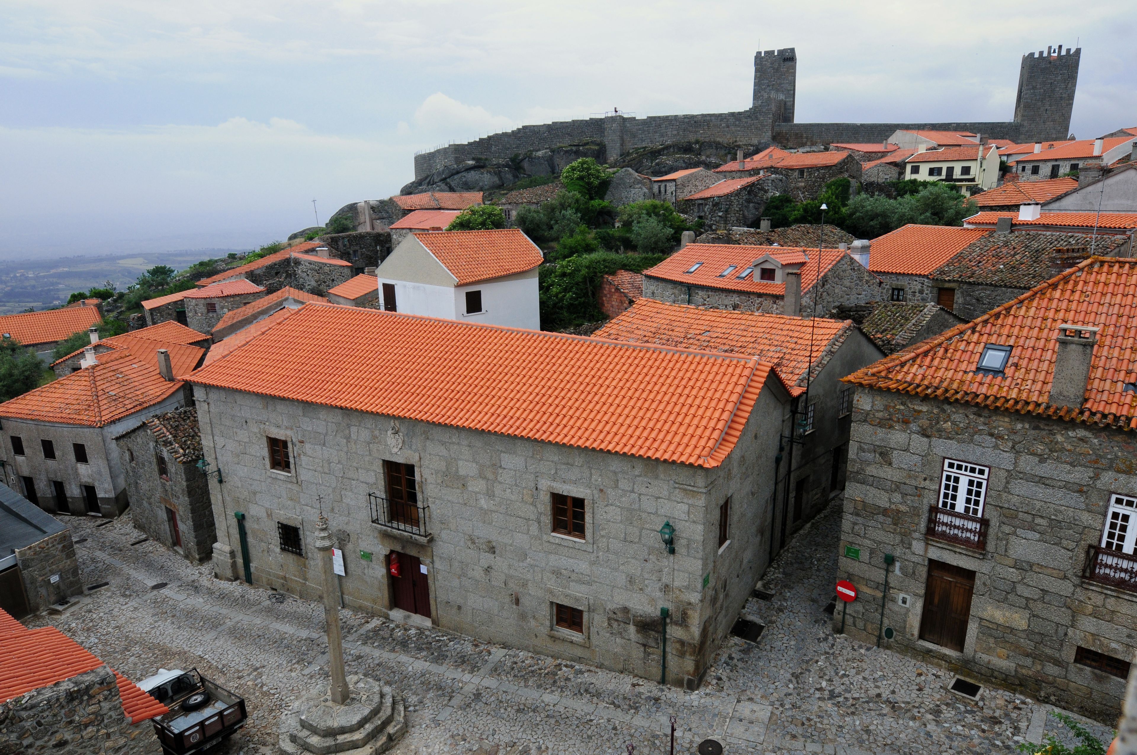 A historic town with stone buildings and red-tiled roofs, a prominent castle in the background, and cobblestone streets.