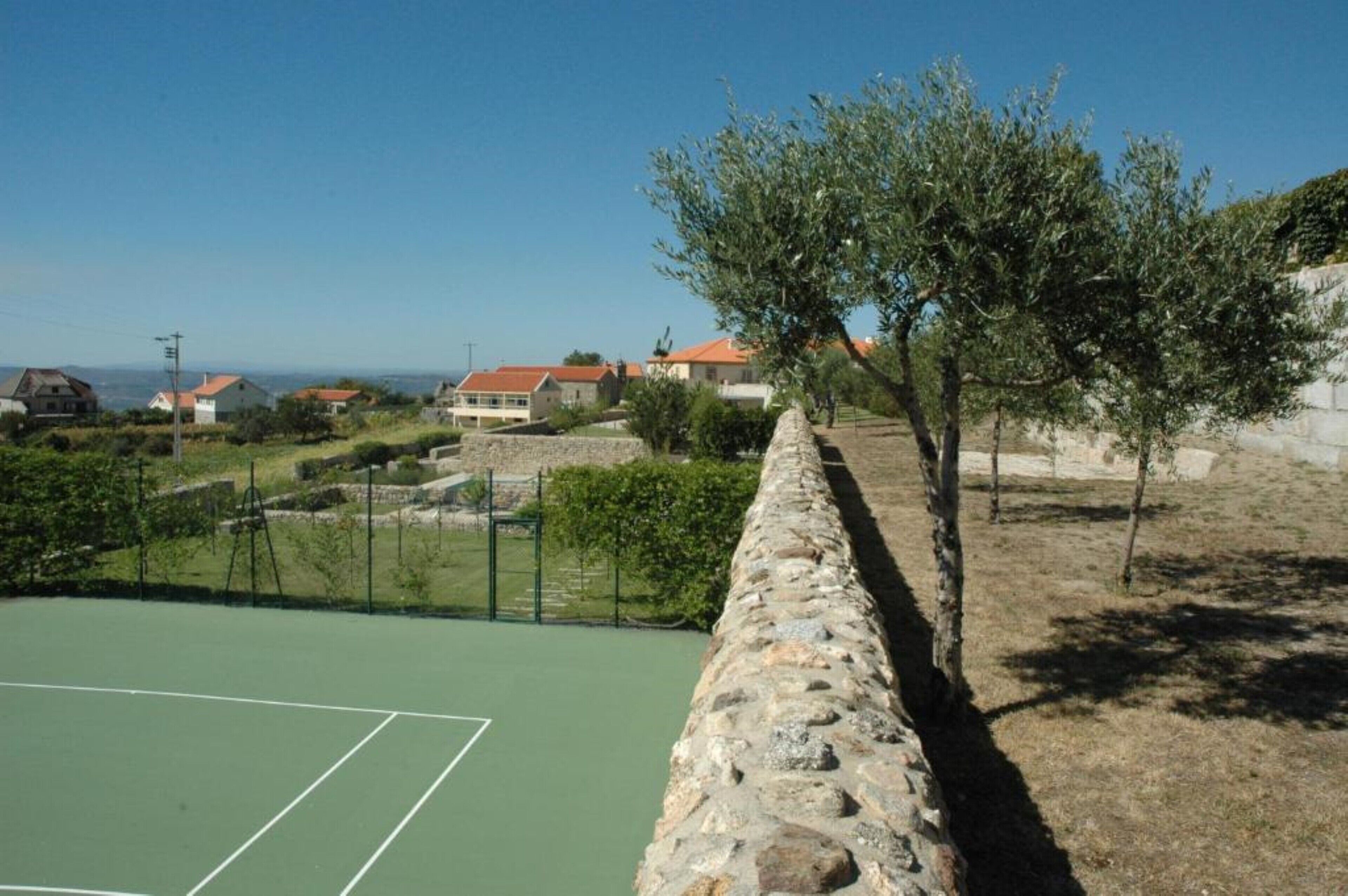 Un court de tennis bordé d’un mur de pierre, avec des oliviers et des maisons en arrière-plan.