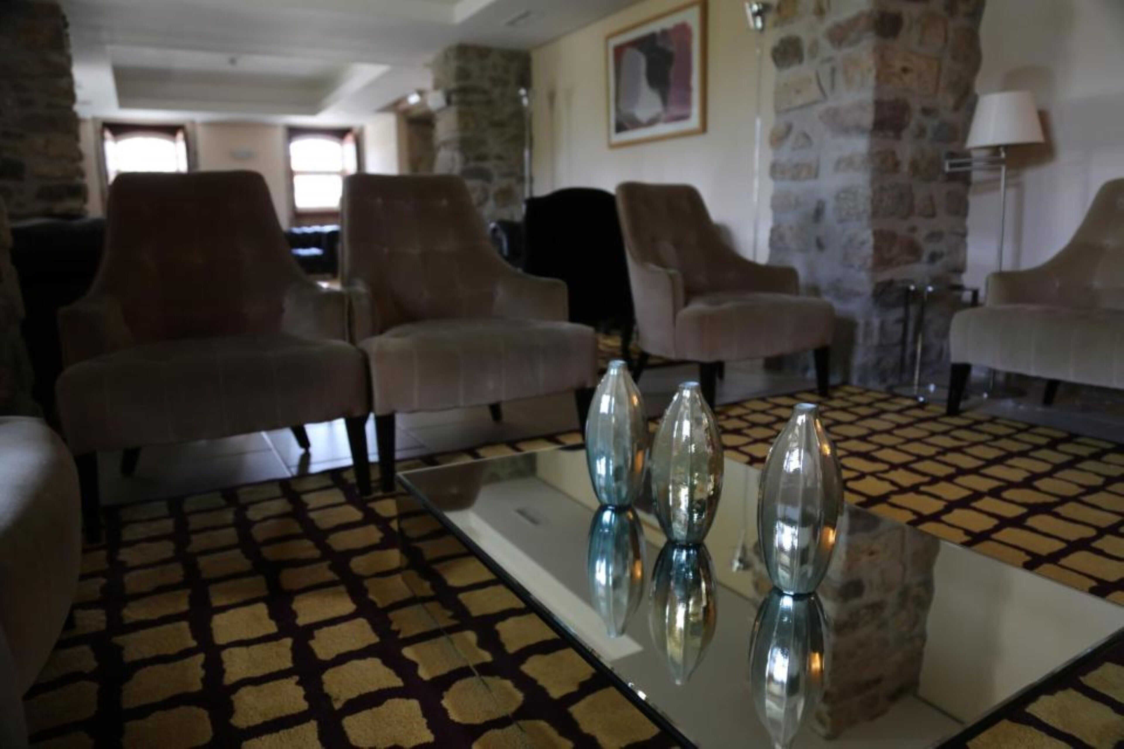 A living room with four armchairs, a glass coffee table with three vases, patterned carpet, and stone walls.