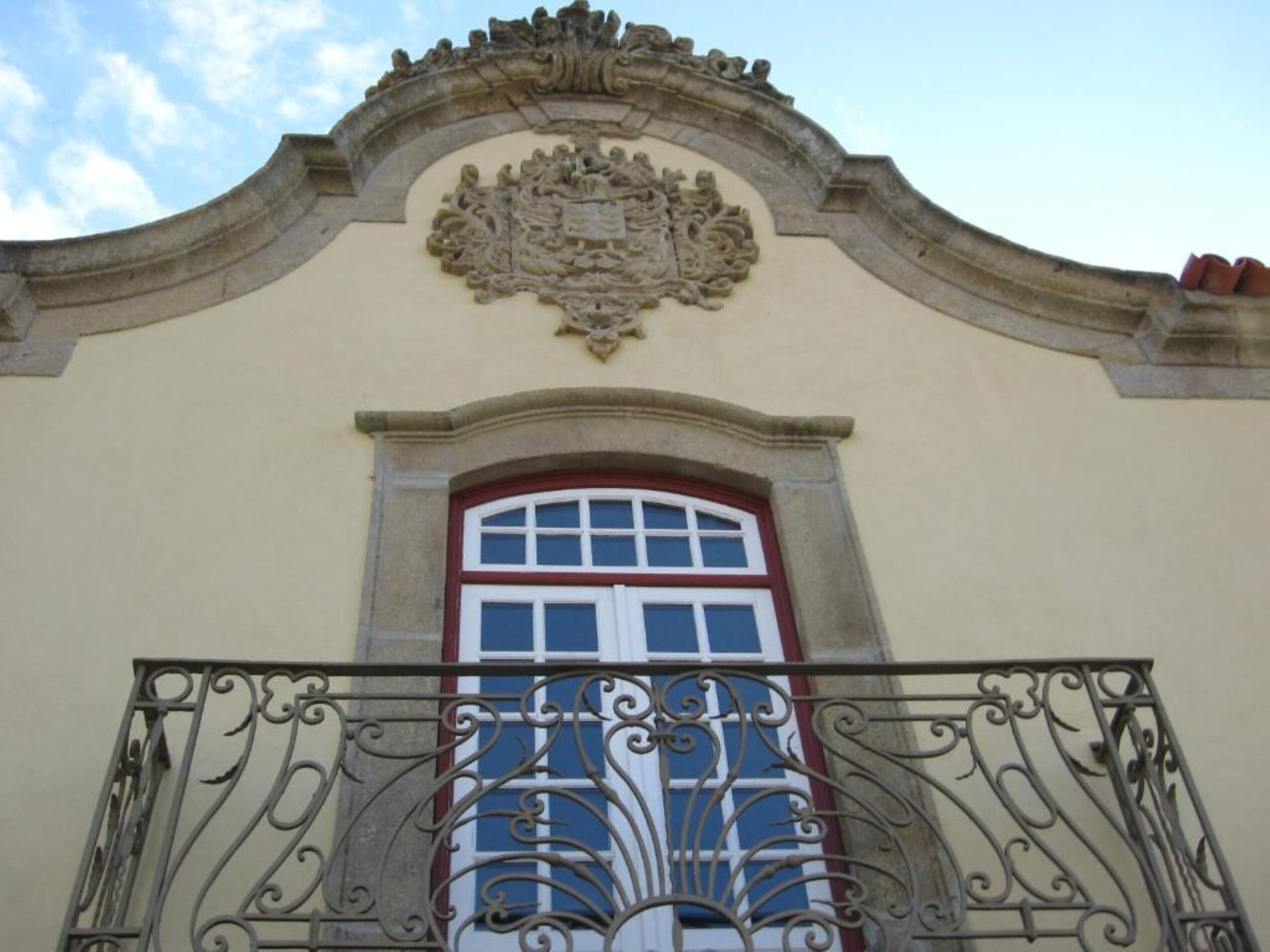 Fachada de edifício com um brasão decorativo sobre uma janela e elaboradas grades de ferro forjado na varanda.
